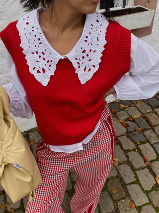 Person wearing a red sweater vest over a white shirt with a lace collar, red checkered pants, and carrying a beige bag.