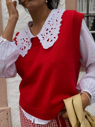 Person wearing a red sweater vest with white lace collar and white long-sleeve shirt.