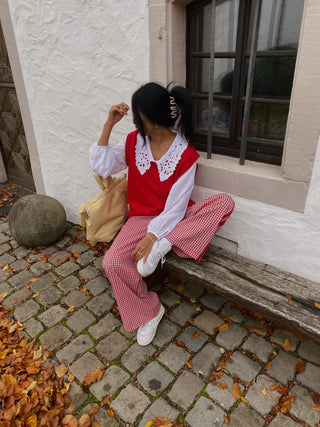 Person sitting on a bench wearing a red vest and pink checkered pants, holding a brown paper bag.