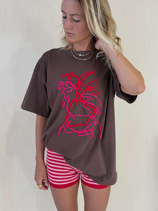 Woman wearing a brown oversized t-shirt with a red graphic design and red and white striped shorts on a plain background.