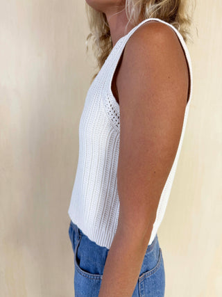 Person wearing a white sleeveless knitted top and blue jeans against a beige background