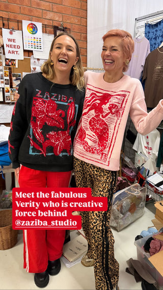 Two women laughing in a clothing boutique. The woman on the left wears a black crewneck sweatshirt with a red graphic print and bright red pants.