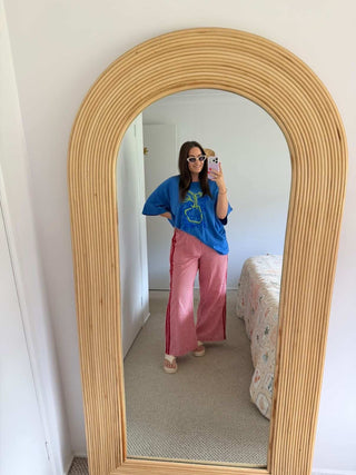 Person taking a mirror selfie wearing a blue top and pink pants in front of a wooden-framed mirror.