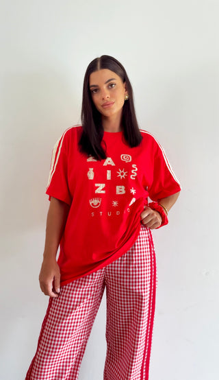 A woman modeling a red graphic t-shirt with white sleeve stripes and matching red-and-white gingham pants, standing against a plain light gray wall.