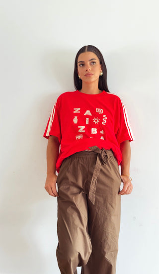 A woman modeling a red short-sleeve graphic t-shirt with white stripes on the sleeves and white text on the front, paired with brown drawstring pants, standing against a plain white wall.