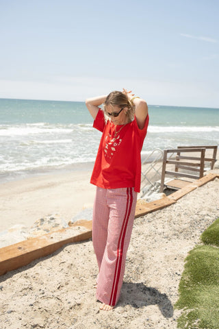 A woman stands on a sandy beach wearing a bright red graphic t-shirt and matching red-and-white gingham pants with red side stripes. The background shows the ocean, a wooden bench, and a clear blue sky.