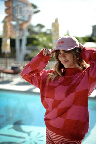 Person wearing a red and pink checkered sweater and pink cap by a poolside.