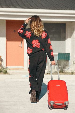 Person wearing a black sweater with red floral patterns and black pants, pulling a red suitcase.