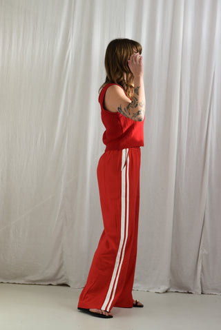 A woman stands in profile wearing a bright red sleeveless top and matching wide-leg pants with white side stripes, posed against a draped white fabric backdrop.