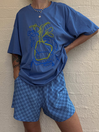 Person wearing a blue oversized t-shirt with a graphic design and matching blue checkered shorts against a beige wall.